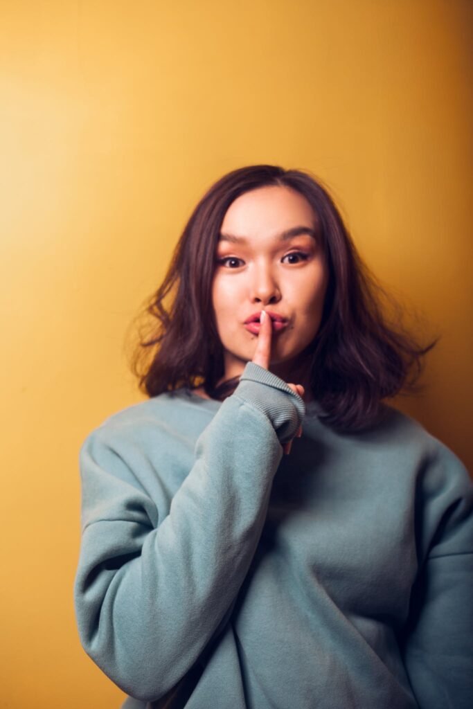 Young Asian female in warm clothing doing shh gesture with finger on lips and looking at camera while standing near orange wall - signifies confidentiality